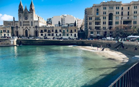 St Julian's Bay with boats on blue water and historic buildings along the shore, under a clear blue sky