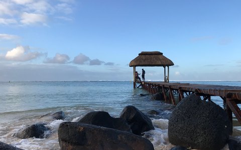 A thatched gazebo at the end of a wooden jetty over the sea at Bel Ombre, with dark rocks in front and white clouds in a blue sky