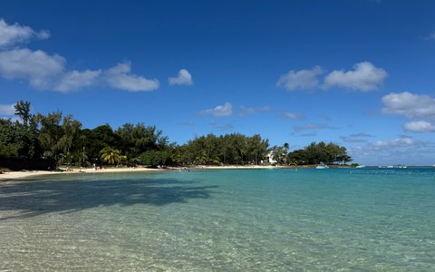 Clear turquoise shallows and a sandy shore lined with trees at Blue Bay, under a blue sky with white clouds