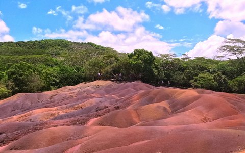 Rippled dunes of red and purple earth at Chamarel in front of green forest, under a blue sky with clouds