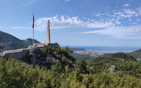 Green hills falling to the sea with the town of Bar along the bay, a stone monument and flag on the hilltop and a blue sky with light cloud