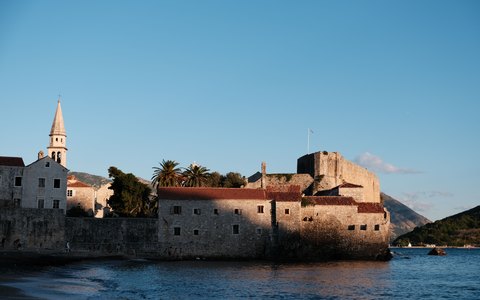 Steinmauern, Glockenturm und rote Dächer der Altstadt von Budva am Wasser, dahinter Berge im warmen Abendlicht