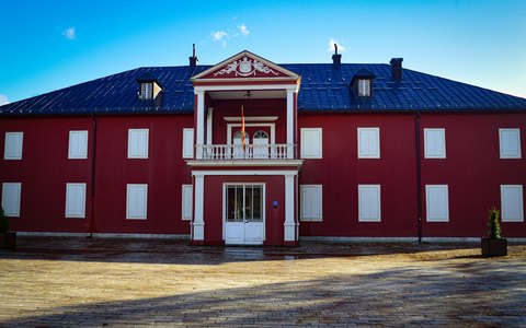 A red two-storey palace with white windows and a balcony, under a blue sky with white clouds