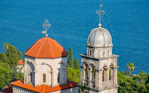 The orange dome and bell tower of a monastery church among green trees, with the blue sea of the bay behind