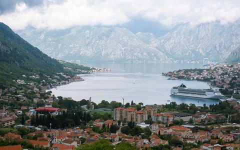 Kotor's roofs below steep grey mountains, with the bay, a moored cruise ship and a cloudy sky
