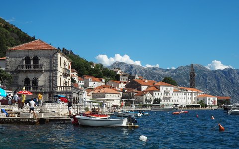 Stone houses and boats along the Perast waterfront, with mountains across the bay and white clouds in a blue sky