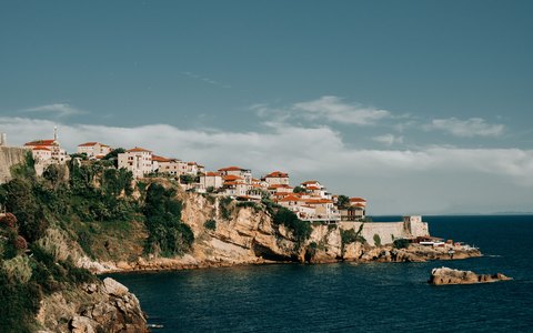 Old stone buildings on a green clifftop above the blue Adriatic, under a pale blue sky