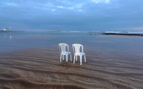 Two white plastic chairs standing in shallow water on a wide flat beach at El Jadida, under a soft evening sky