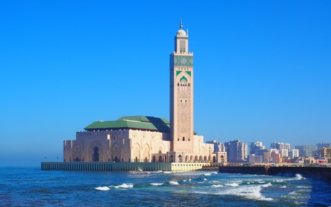 The Hassan II Mosque and its tall minaret on the shore at Casablanca, with ocean waves and a vivid blue sky
