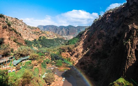 A green canyon with red cliffs and a rainbow in the spray of the Ouzoud falls, under a clear blue sky