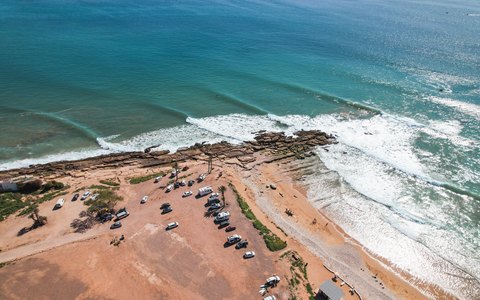 Aerial view of the Taghazout shore with waves breaking on a sandy point and vans parked on the cliff top