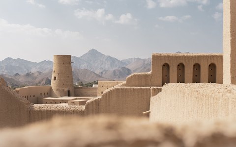The mud-brick walls and towers of Bahla Fort with the Hajar Mountains behind, under a pale sky with light cloud