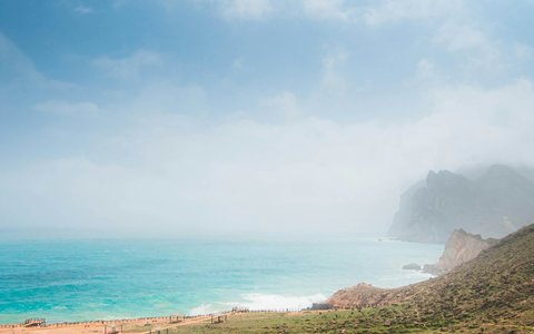 Turquoise sea and a curving beach at Mughsail, with mist over the coastal cliffs and green slopes in the foreground