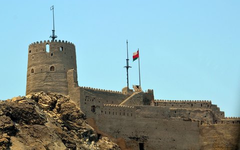 The stone Muttrah Fort with its round tower and flag on a rocky hill, under a clear blue sky