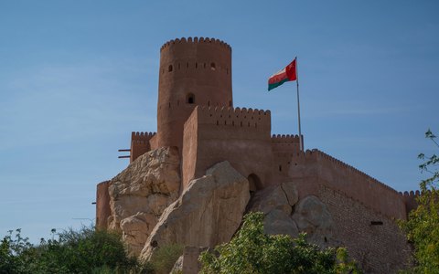 The round stone tower of Nakhal Fort with the Omani flag on a rocky hilltop, under a deep blue sky