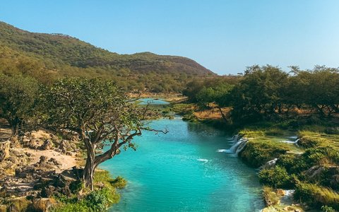 A turquoise stream with small waterfalls running between green trees and rocks, below wooded hills and a blue sky