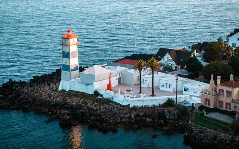 Aerial view of the Santa Marta lighthouse and white buildings on the rocky shore at Cascais, beside the blue sea