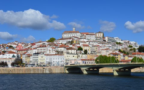 Coimbra's old town and university tower rising above the Mondego River and a bridge, under a blue sky with white clouds