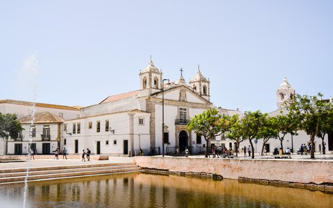 White church with twin bell towers beside a square and a shallow pool in Faro