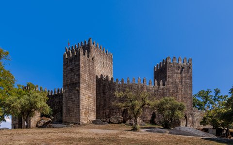 The stone keep and battlements of Guimaraes Castle among trees, against a clear blue sky