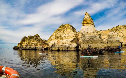 Golden sandstone sea stacks and cliffs at Lagos reflected in calm green-blue water, with paddleboarders and a cloudy blue sky
