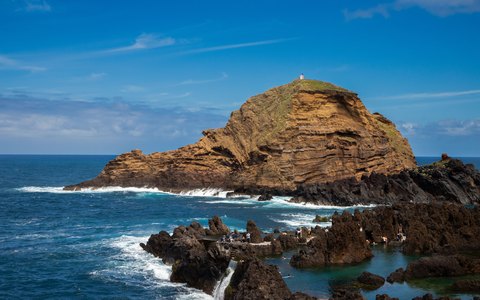 A rocky islet topped by a small lighthouse off the lava coast of Porto Moniz, with waves and a blue sky