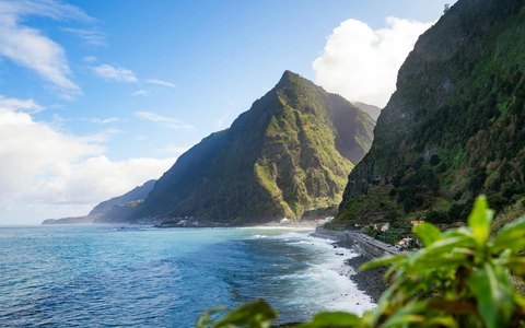 Steep green mountains dropping to a rocky shore and blue sea near Sao Vicente on Madeira's north coast, under a blue sky with white clouds