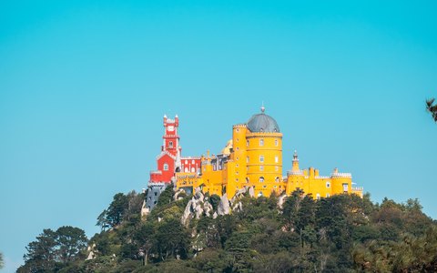 The red and yellow towers of Pena Palace on a wooded hilltop against a clear blue sky