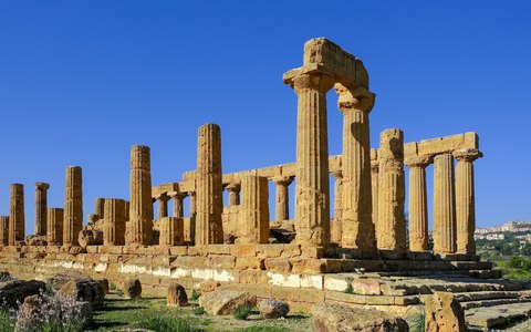 A Doric temple of golden stone with rows of columns on a grassy ridge, under a deep blue sky