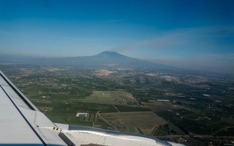 Flugzeugflügel über grünen und braunen Feldern in der Ebene bei Catania, am Horizont der Ätna mit Rauchfahne