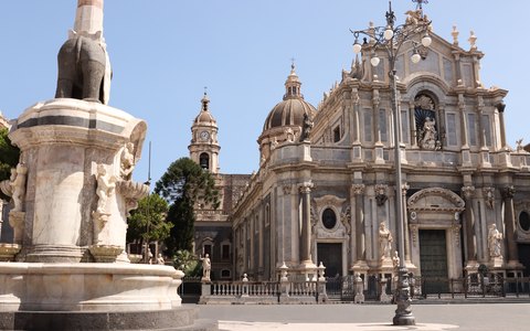 The baroque facade of Catania Cathedral beside the elephant fountain in Piazza Duomo, under a blue sky with light cloud