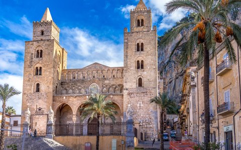 The twin-towered Norman cathedral of Cefalu with palm trees in front, under a bright blue sky