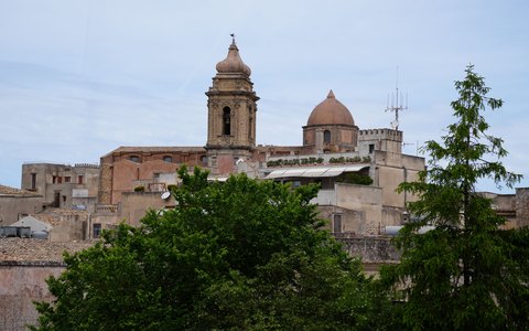 An old stone church tower and rooftops in Erice behind green trees, under a blue sky with white clouds