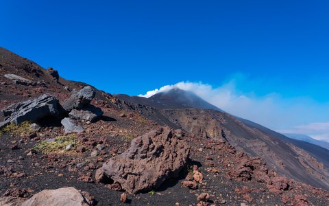 Dark volcanic slopes of Mount Etna with a smoking summit against a deep blue sky