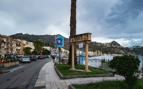 A seafront road at Giardini Naxos with palm trees and a stone column, with the hills of Taormina behind under a cloudy sky