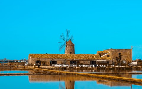 A stone windmill and salt works reflected in still water under a bright blue sky
