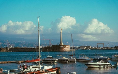 Vertäute Boote im Hafen von Messina mit der hohen Statuensäule an der Hafenspitze unter blauem Himmel mit weißen Wolken