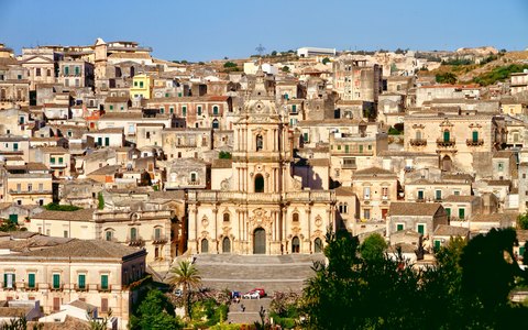 The baroque cathedral of San Giorgio rising above the stone houses of Modica, under a blue sky