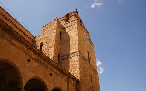 The golden stone bell tower of Monreale Cathedral seen from below against a vivid blue sky