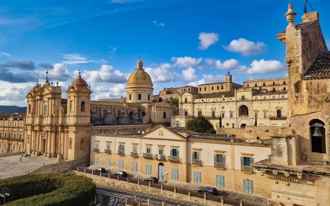 The baroque Noto Cathedral and nearby palaces in warm stone, under a blue sky with white clouds