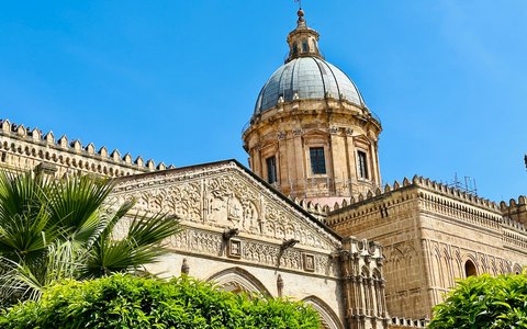 The ornate facade and dome of Palermo Cathedral above palm trees, under a clear blue sky