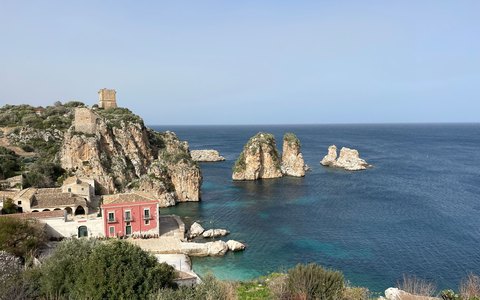 Sea stacks and a tower on the Scopello shore above clear blue water, under a blue sky