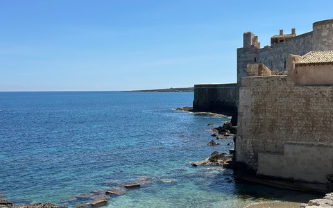 Old stone buildings of Ortigia rising straight from the clear blue sea under a blue sky