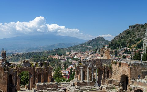 The brick ruins of the ancient theatre at Taormina with the town, the coast and Mount Etna beyond under a blue sky