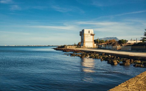 An old stone tower on the Trapani shore at the end of a rocky breakwater, with calm sea and soft evening light