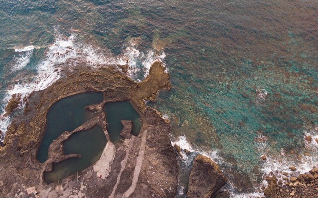 Aerial view of dark rock pools on a rocky point at Agaete, with waves breaking along the shore and green-blue sea around