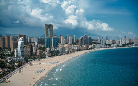 Ein langer Sandstrand, der sich unterhalb der Hochhäuser von Benidorm in einem Bogen erstreckt, mit türkisfarbenem Meer und Wolken