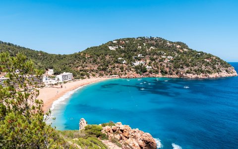 Sandbucht mit türkisfarbenem Wasser unterhalb eines grünen Hügels bei Cala Sant Vicent auf Ibiza