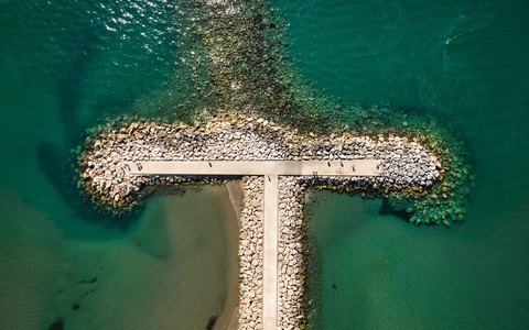 Aerial view of a T-shaped stone breakwater reaching into clear green-blue sea