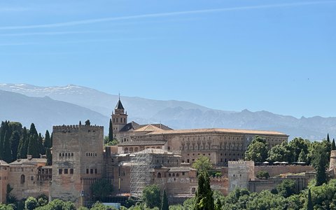 The walls and towers of the Alhambra above green trees, with the Sierra Nevada mountains behind under a blue sky